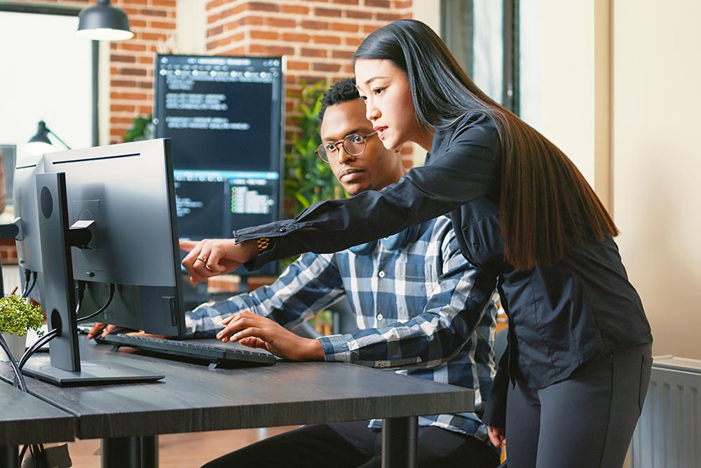Two programers holding laptop with coding interface walking towards desk and sitting down talking about online cloud computing. Software developers team discussing algorithms on computer screen.