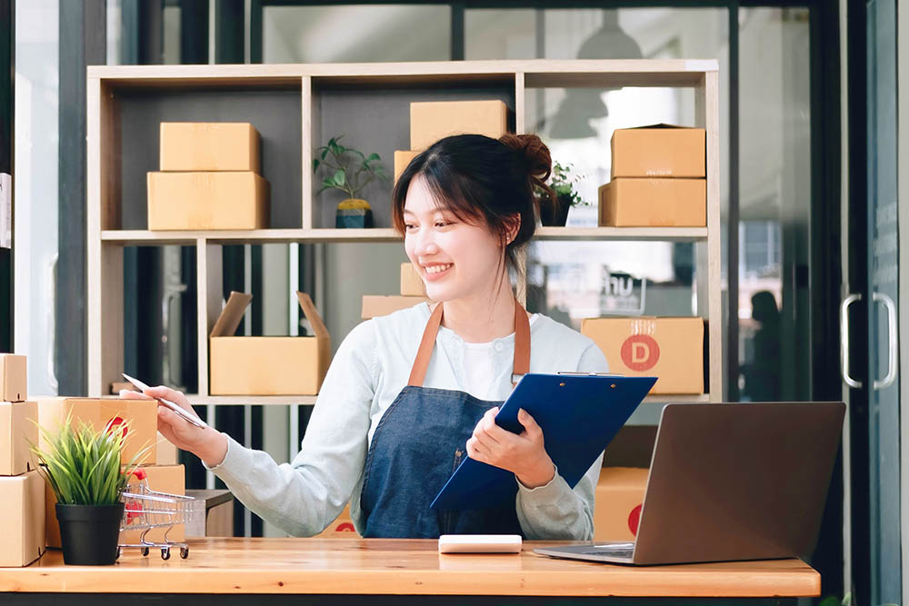 A portrait of a young Asian woman, e-commerce employee sitting in the office full of packages in the background write note of orders and a calculator, for SME business, e-commerce and delivery business.